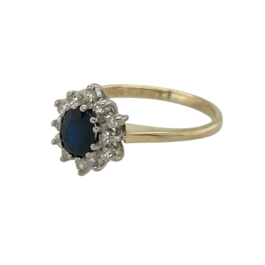 Bring classic sparkle and rich colour to your collection with this pre-owned 9ct yellow gold sapphire and diamond cluster ring—an elegant piece offering timeless beauty and refined charm.
Design: This size J ring features a 5mm by 4mm oval sapphire at the centre, surrounded by a halo of diamonds that enhance its deep blue tone with bright, eye-catching sparkle.