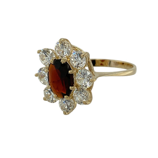 Bring rich colour and classic sparkle to your jewellery collection with this pre-owned 9ct yellow gold garnet & cubic zirconia set flower cluster ring, a beautifully balanced design with timeless appeal.
Design: The ring features a floral-inspired cluster arrangement, centred with an oval garnet measuring 8mm by 6mm and surrounded by cubic zirconia stones, each measuring approximately 4mm in diameter, creating a radiant and eye-catching focal point.