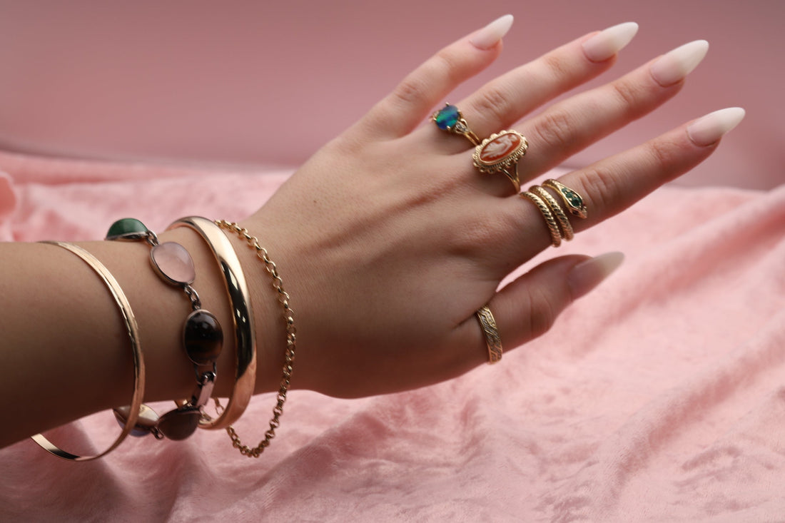 A hand with stacked gold and silver pre-owned vintage jewellery including a bracelet, bangles, and four gemstone set cameo snake rings