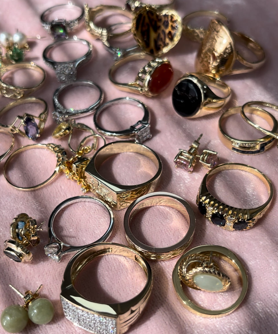 Range of gold rings and stud earrings on a pink background. The jewellery is set with mixed gemstones and diamonds