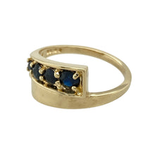 Load image into Gallery viewer, Add graceful colour to your jewellery collection with this pre-owned 9ct yellow gold and sapphire set crossover ring, featuring:

Design: A stylish crossover band set with four sapphires in graduating size, creating a flowing and elegant look.
Material: 9-carat yellow gold, offering a warm, polished finish that complements the rich blue tones of the stones.
Ring Front: Measures 9mm wide at the front, making a bold yet refined statement.
Gemstone Detail: Set with four sapphires in a graduated arrangement
