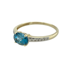 Load image into Gallery viewer, Bring vibrant colour and refined sparkle to your collection with this pre-owned 9ct yellow gold ring, featuring a striking blue topaz and shimmering diamond accents—perfect for elegant everyday wear or special occasions.

Design: The ring showcases a 6mm by 8mm oval-cut blue topaz as its centrepiece, set horizontally for a modern twist. The band is half-set with brilliant-cut diamonds, adding a touch of sparkle that enhances the vivid blue of the topaz.

