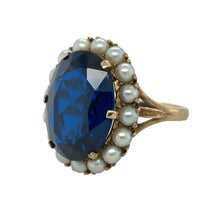 Load image into Gallery viewer, Bring vintage-inspired elegance and striking colour to your collection with this pre-owned 9ct yellow gold dress ring, featuring a bold oval blue stone framed by delicate seed pearls.

Design: The ring showcases a 16mm by 12mm oval-shaped blue stone surrounded by a halo of seed pearls, creating a refined and eye-catching design.
Material: Made from 9-carat yellow gold, the polished finish enhances the warm tone of the gold and complements the rich blue centre and soft lustre of the pearls.
