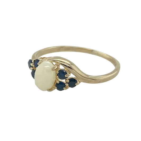 Bring elegant colour and timeless charm to your collection with this pre-owned 9ct yellow gold sapphire and opalique ring, a delicate design with classic appeal.

Design: The ring features a 6mm by 4mm oval opalique stone at the centre, accented on each side by three deep blue sapphires for a refined and balanced look.
Material: Made from 9-carat yellow gold, the polished finish enhances the play of colour in the opalique and the richness of the sapphires.