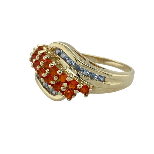 Bring vibrant colour and artistic flair to your collection with this pre-owned 9ct yellow gold orange stone and tanzanite wave band ring—an expressive piece that combines flowing design with striking contrast.

Design: This eye-catching ring features a wave-style band set with a vivid orange stone and complementary tanzanite accents, creating a bold interplay of warm and cool tones. The front of the band measures approximately 11mm wide, giving it a strong and stylish presence on the finger.