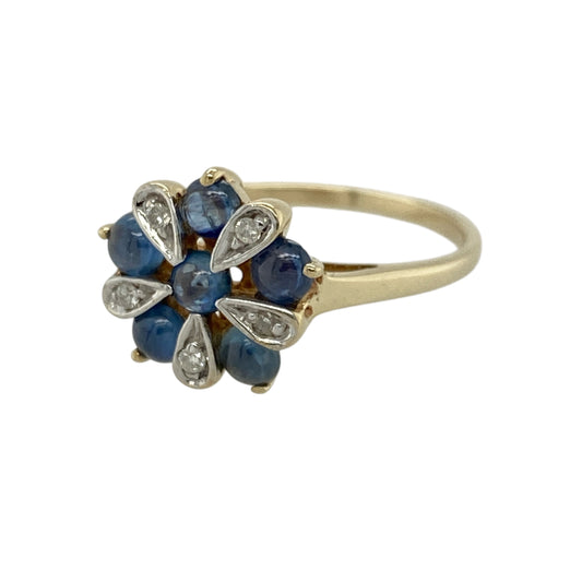 Add elegant sparkle and floral-inspired charm to your jewellery collection with this pre-owned 9ct yellow gold diamond and sapphire flower cluster ring, featuring a bold and eye-catching design—perfect for special occasions or standout everyday wear.

Design: The ring is composed of multiple sapphires arranged to form a flower, with white gold-set diamonds creating petal detailing that adds contrast and brilliance.