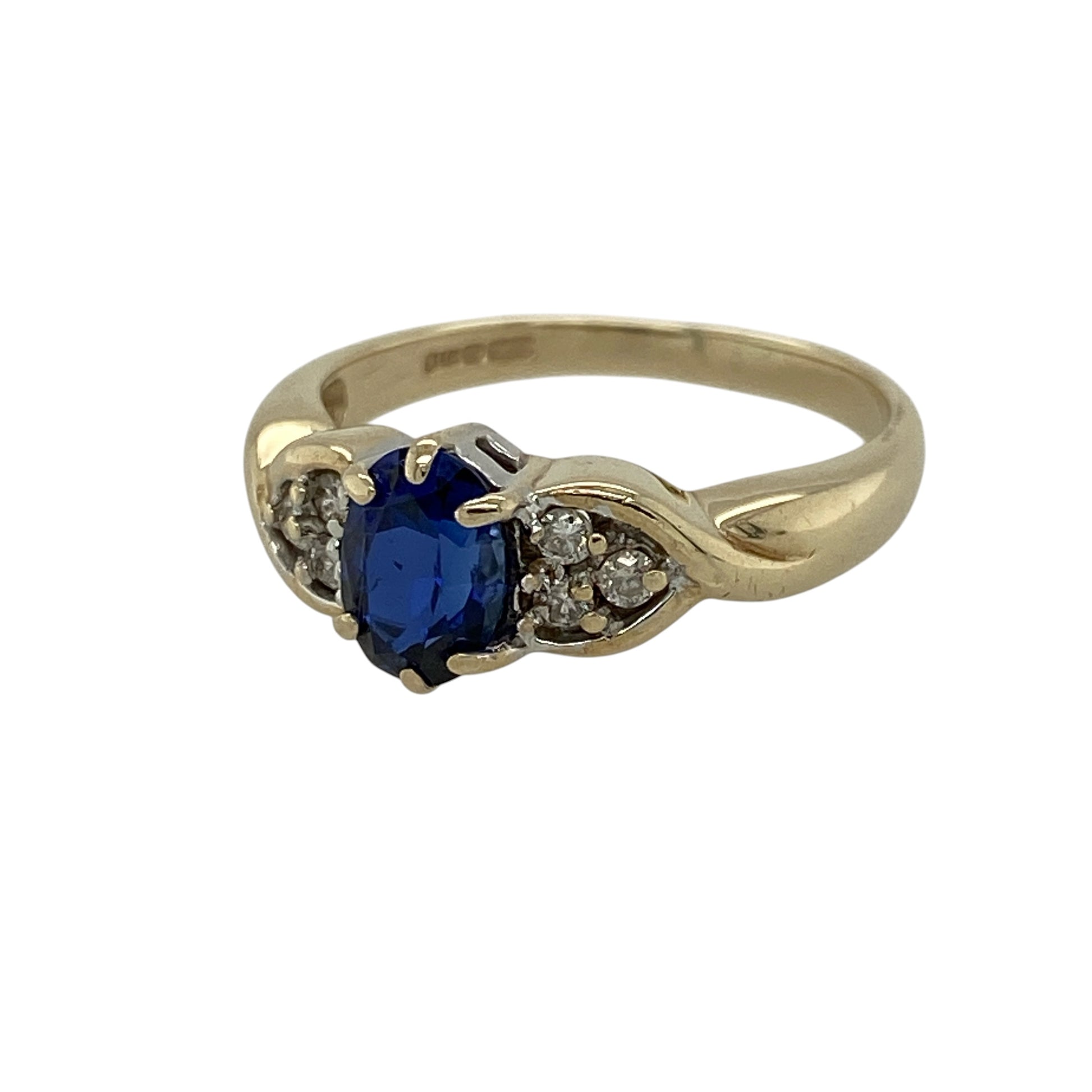 Add elegant colour and flowing sparkle to your jewellery collection with this pre-owned 9ct yellow gold diamond and sapphire-coloured stone ring, featuring a graceful design and eye-catching detail—perfect for everyday wear or special occasions.

Design: The ring showcases an oval sapphire-coloured stone measuring approximately 7mm by 5mm at its centre, complemented by diamond set twist shoulders that add brilliance and movement.