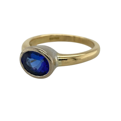 Bring bold colour and refined elegance to your collection with this pre-owned 18ct yellow gold blue stone solitaire ring, featuring a striking centrepiece with contemporary detailing—perfect for special occasions or standout everyday wear.

Design: The ring showcases a 9mm by 7mm oval blue stone at the centre, presented in a white gold rubover style for a smooth, modern finish and a clean, defined look.